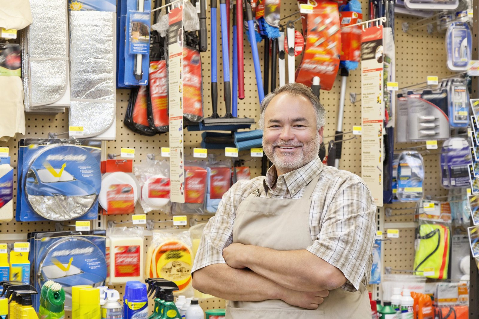 Smiling man in an apron standing in a small business retail setting, surrounded by various cleaning and automotive supplies, representing local business marketing and customer engagement.