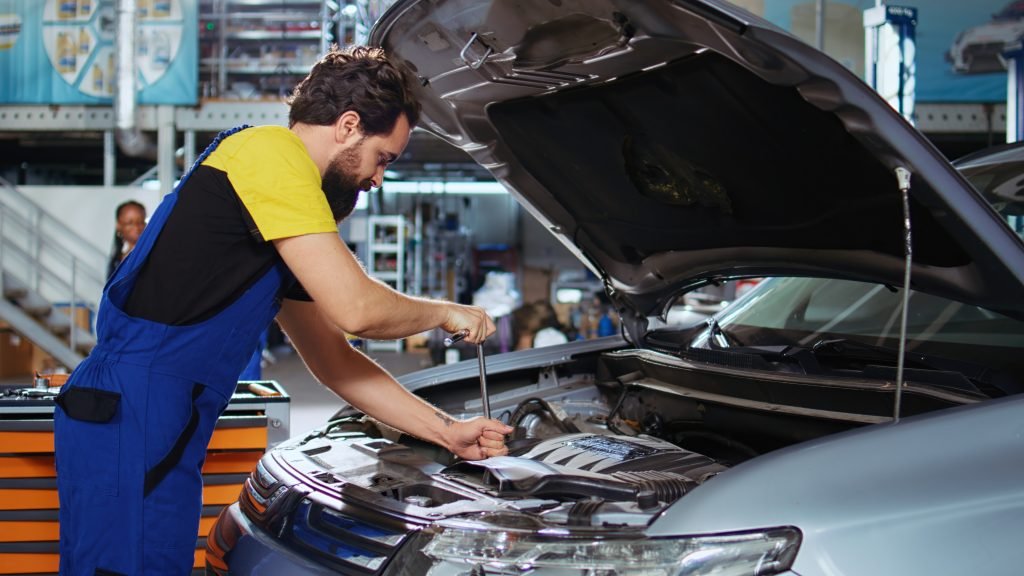 Car technician in blue overalls working on vehicle engine in auto repair shop, emphasizing hands-on service and expertise in automotive maintenance.
