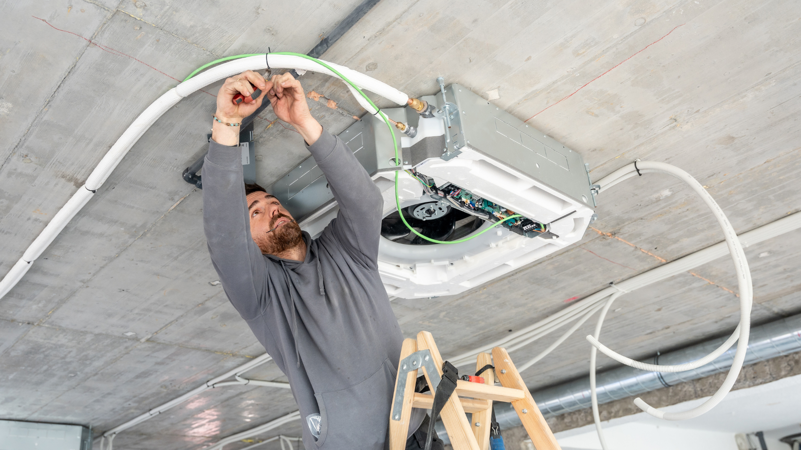 Electrician working on HVAC installation, adjusting wiring and components on ceiling, with ladder and cables visible.