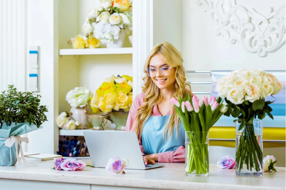 Woman in a flower shop smiling while working on a laptop, surrounded by fresh flowers and greenery, illustrating efficient service and digital marketing for local businesses.