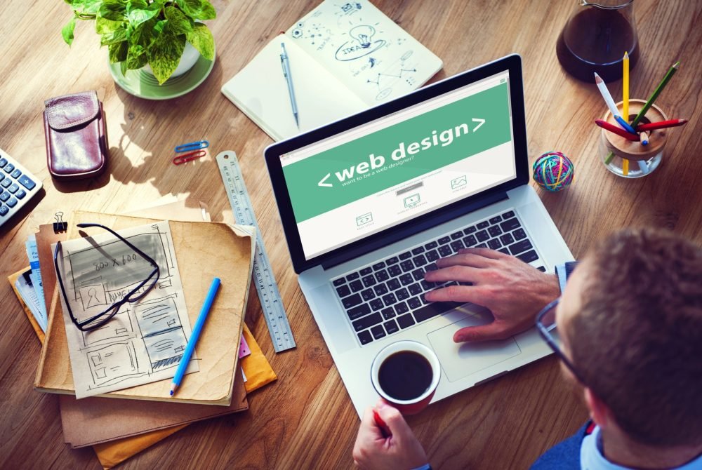 Person working on laptop displaying web design, surrounded by stationery, sketches, and a coffee cup, emphasizing digital marketing and website development.