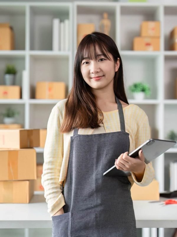 Young woman entrepreneur in home office, holding digital tablet, surrounded by shipping boxes, representing AM Marketing Group's focus on local business growth.