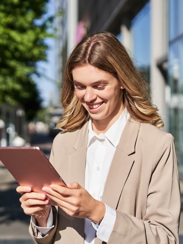 Happy businesswoman smiling while using a digital tablet outdoors, near office buildings, reflecting AM Marketing Group's focus on digital marketing solutions for local businesses.